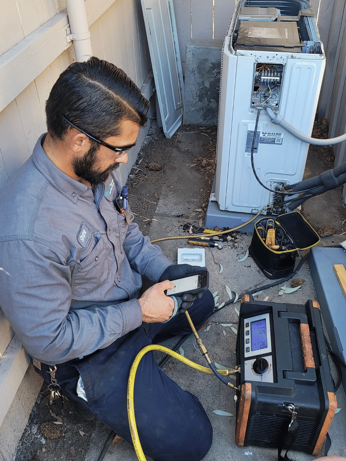 A technician kneeling at an outdoor condenser, running a live measureQuick diagnostic on a phone with the analyzer kit beside him — the kind of call a remote coach can join.