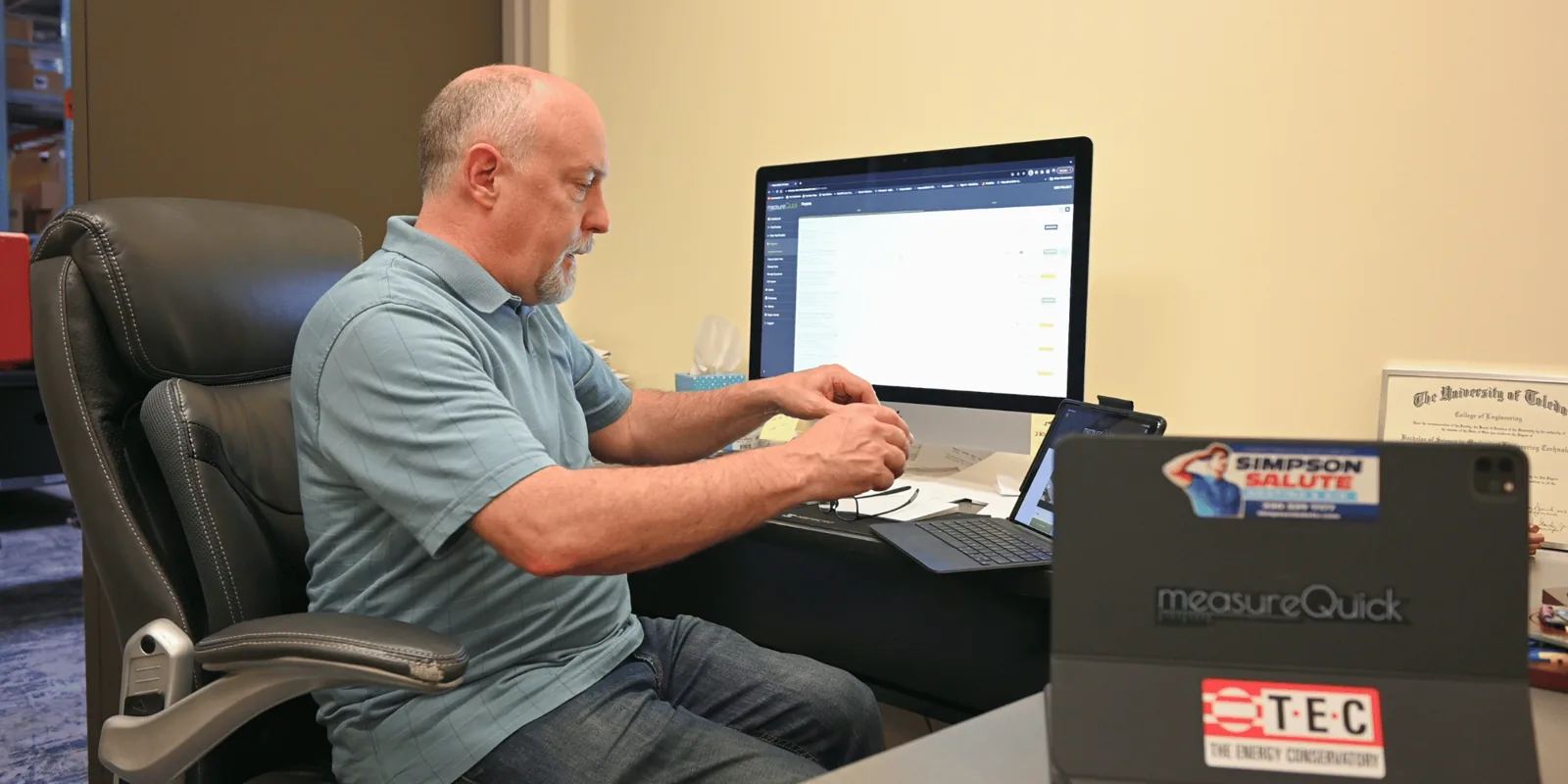 Manager at an office desk reviewing the measureQuick cloud dashboard on a monitor, tablet alongside