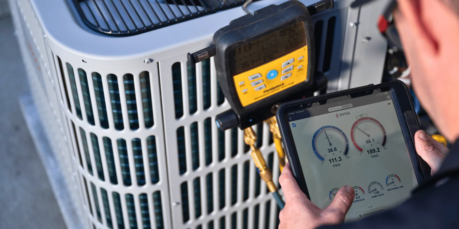 Technician at an outdoor condenser reading measureQuick on a tablet, manifold connected to the service ports