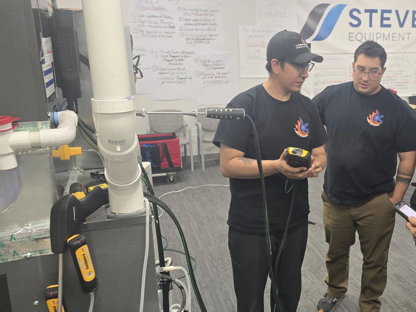 HVAC students gathered around a lab training rig, one reading live measurements off a handheld instrument