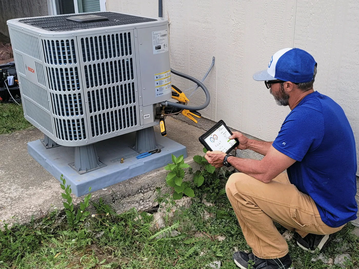 HVAC technician kneeling at an outdoor condenser, reading measureQuick on a tablet with wireless probes clipped to the service lines